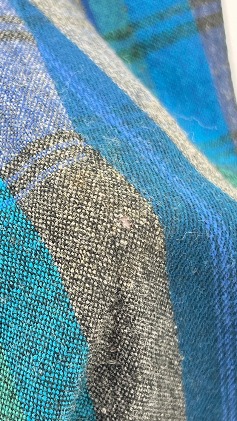 May include: Close-up of a woven fabric with a plaid pattern. The design features a combination of blue, teal, gray, and white hues. The texture of the fabric appears soft and slightly fuzzy, with visible threads.