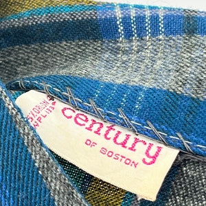 May include: Close-up of a blue, gray, and yellow plaid fabric with a white tag. The tag has pink text that reads "century OF BOSTON". The fabric is stitched with white thread.