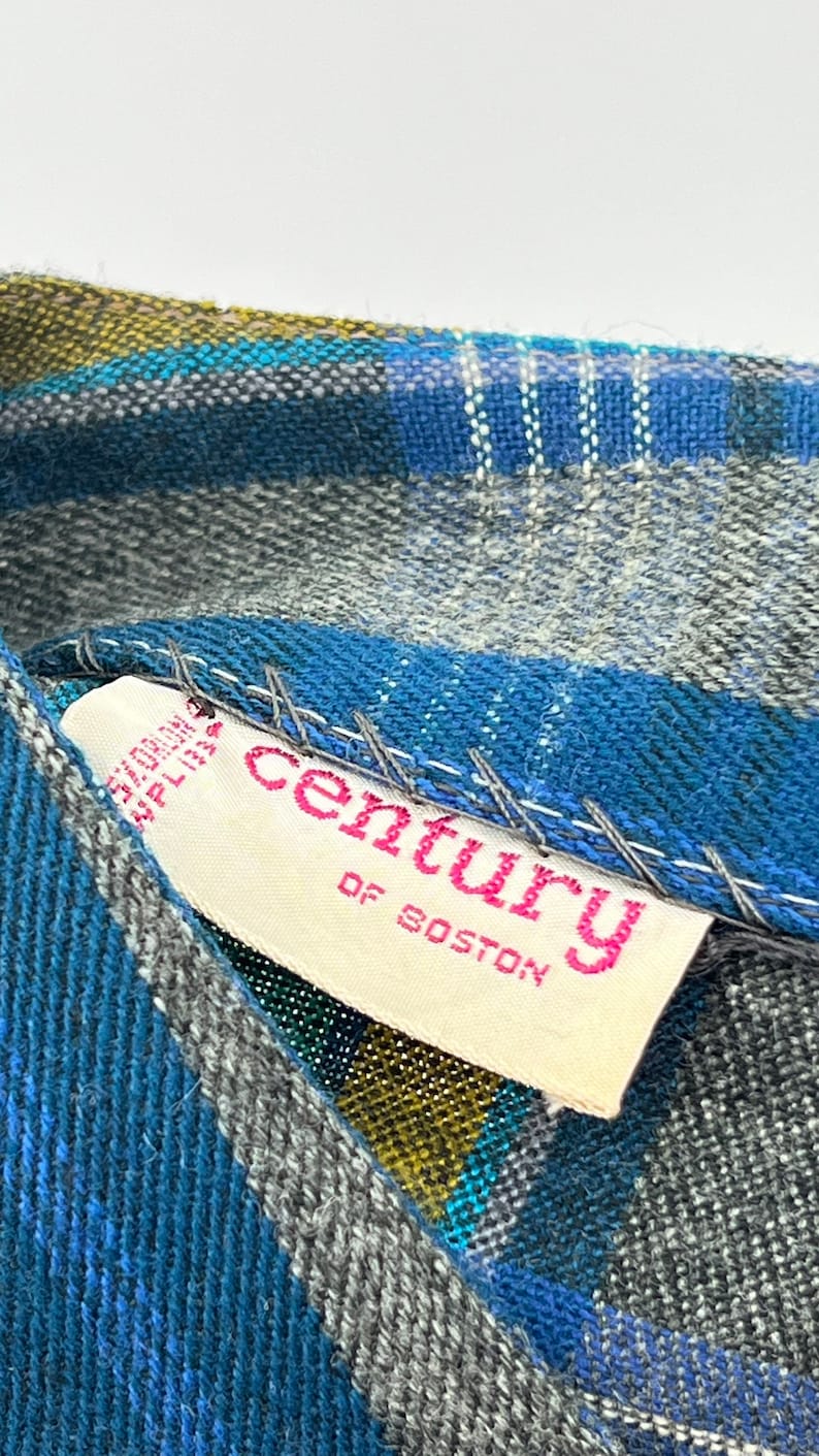 May include: Close-up of a blue, gray, and yellow plaid fabric with a white tag. The tag has pink text that reads "century OF BOSTON". The fabric is stitched with white thread.