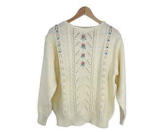 May include: A white knitted sweater with a delicate openwork design and floral embroidery. The sweater has a crew neck and long sleeves.