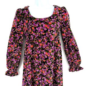 May include: A long-sleeved floral dress with a scoop neckline. The dress features a black background covered in small purple, red, and green flowers. The sleeves have a ruffled cuff. The dress is displayed on a white mannequin.