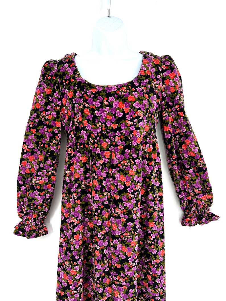 May include: A long-sleeved floral dress with a scoop neckline. The dress features a black background covered in small purple, red, and green flowers. The sleeves have a ruffled cuff. The dress is displayed on a white mannequin.