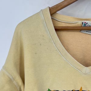 Op de afbeelding: Een lichtgeel V-hals sweatshirt met een vervaagde grafische print. Het sweatshirt heeft een geribbelde hals en manchetten. Het label op het sweatshirt luidt "One Size Fits All".