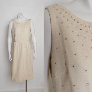 May include: Cream-colored sleeveless dress with a scoop neckline and knee-length hem. The dress is adorned with scattered, sparkling embellishments. The dress is displayed on a mannequin. The dress has a fitted waist and a straight skirt.
