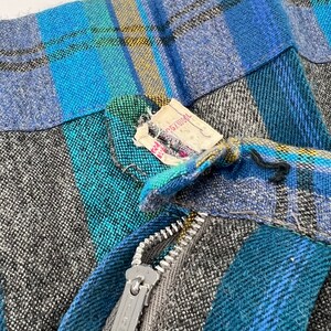 May include: Close-up of a vintage skirt with a plaid pattern in shades of blue, teal, gray, and yellow. The skirt has a silver zipper and a tag attached. The fabric appears to be a woven wool blend.