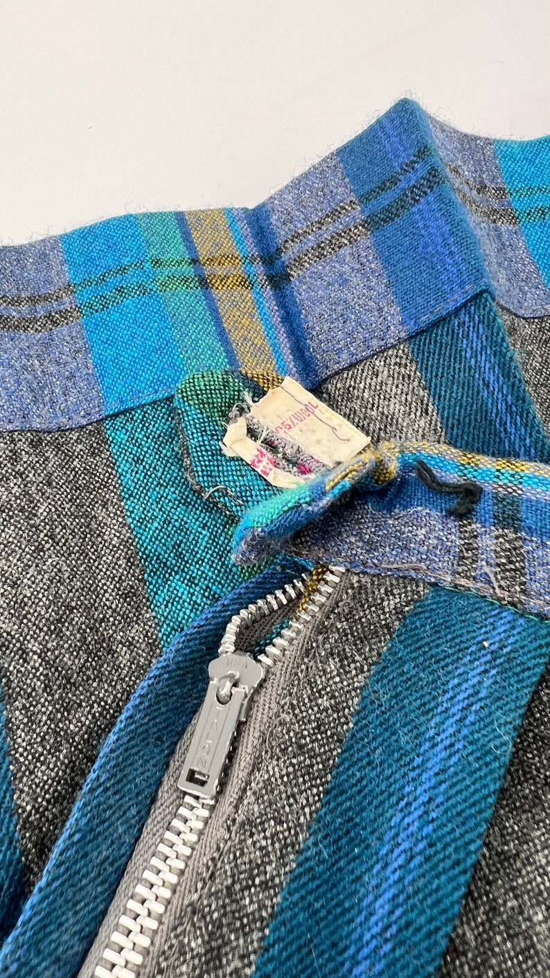 May include: Close-up of a vintage skirt with a plaid pattern in shades of blue, teal, gray, and yellow. The skirt has a silver zipper and a tag attached. The fabric appears to be a woven wool blend.