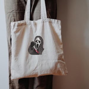 May include: A cream tote bag with a graphic of the Ghostface mask from the Scream horror film series. The design includes a black hooded cloak and a bloody knife. The bag is held by a person.