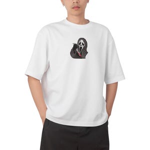 May include: White t-shirt with a black and red graphic of the Ghostface mask holding a bloody knife. The design is centered. The shirt has short sleeves and a relaxed fit.