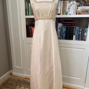 以下が含まれることがあります： マネキンに着せられた、アイボリー色のヴィンテージイブニングドレス。胴部分は小さなパールとゴールドのトリムで装飾されています。ドレスはスクエアネックラインと、流れるようなロングスカートが特徴です。本棚の前で展示されています。