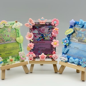Könnte beinhalten: Drei dekorative Pokemon-Kartenrahmen auf kleinen Holzstaffeleien. Die Rahmen sind mit bunten, cartoonartigen Charms verziert, darunter Blumen, Schleifen und Figuren. Jeder Rahmen enthält eine andere Pokemon-Karte, deren Details sichtbar sind.