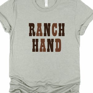 May include: A light gray t-shirt with the words "RANCH HAND" printed in brown and dark brown block letters. The shirt has short sleeves and a crew neck. The shirt is made of a soft material.