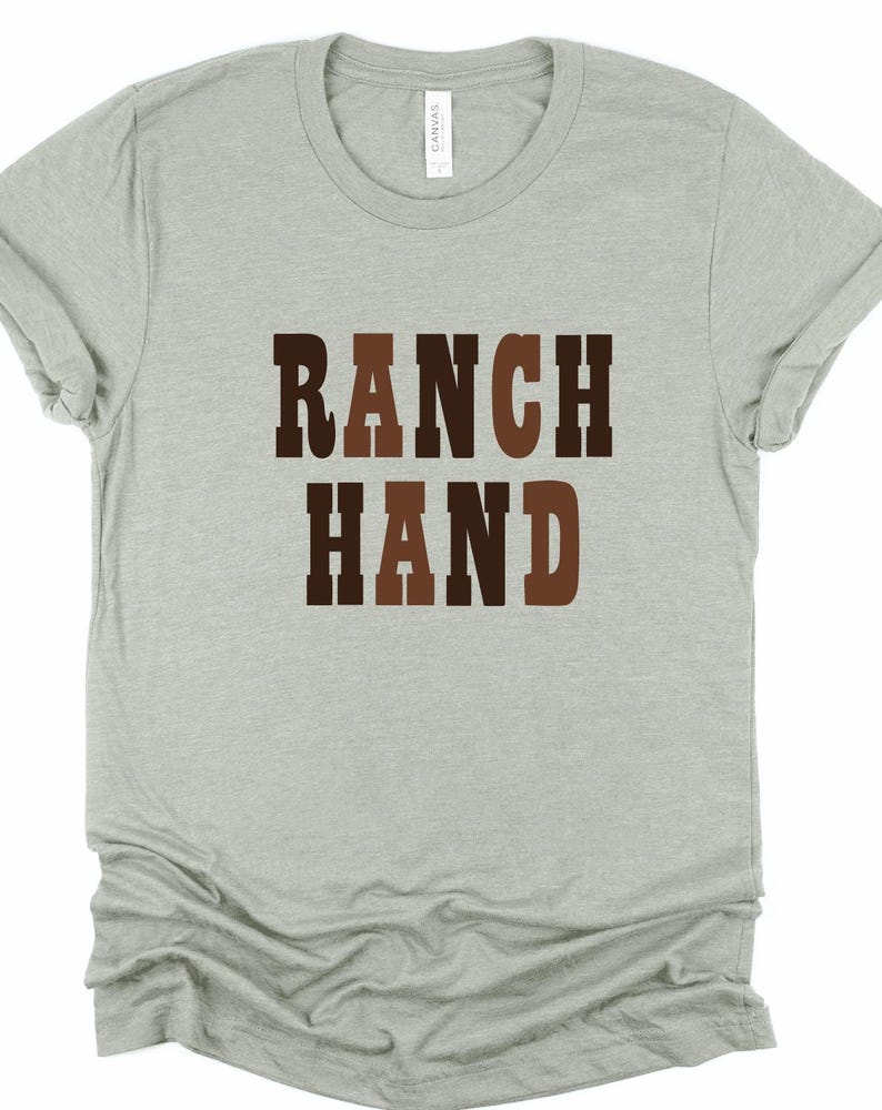 May include: A light gray t-shirt with the words "RANCH HAND" printed in brown and dark brown block letters. The shirt has short sleeves and a crew neck. The shirt is made of a soft material.