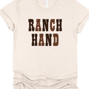 May include: A light beige t-shirt with the words "RANCH HAND" printed in a brown, western-style font. The shirt has short sleeves and a crew neck. The fabric appears to be soft and comfortable.