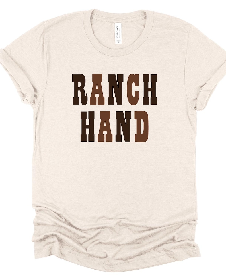May include: A light beige t-shirt with the words "RANCH HAND" printed in a brown, western-style font. The shirt has short sleeves and a crew neck. The fabric appears to be soft and comfortable.