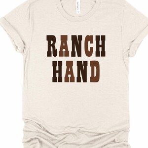 May include: A light beige t-shirt with the words "RANCH HAND" printed in brown and dark brown block letters. The shirt has short sleeves and a crew neck. The fabric appears to be soft and comfortable.