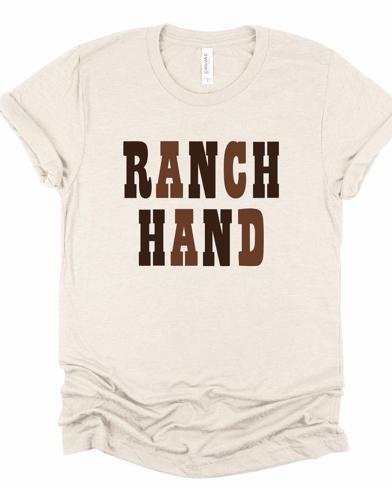 May include: A light beige t-shirt with the words "RANCH HAND" printed in brown and dark brown block letters. The shirt has short sleeves and a crew neck. The fabric appears to be soft and comfortable.