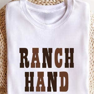 May include: A white t-shirt with the words "RANCH HAND" printed in brown and dark brown block letters. The shirt is folded and resting on a woven, light brown surface.
