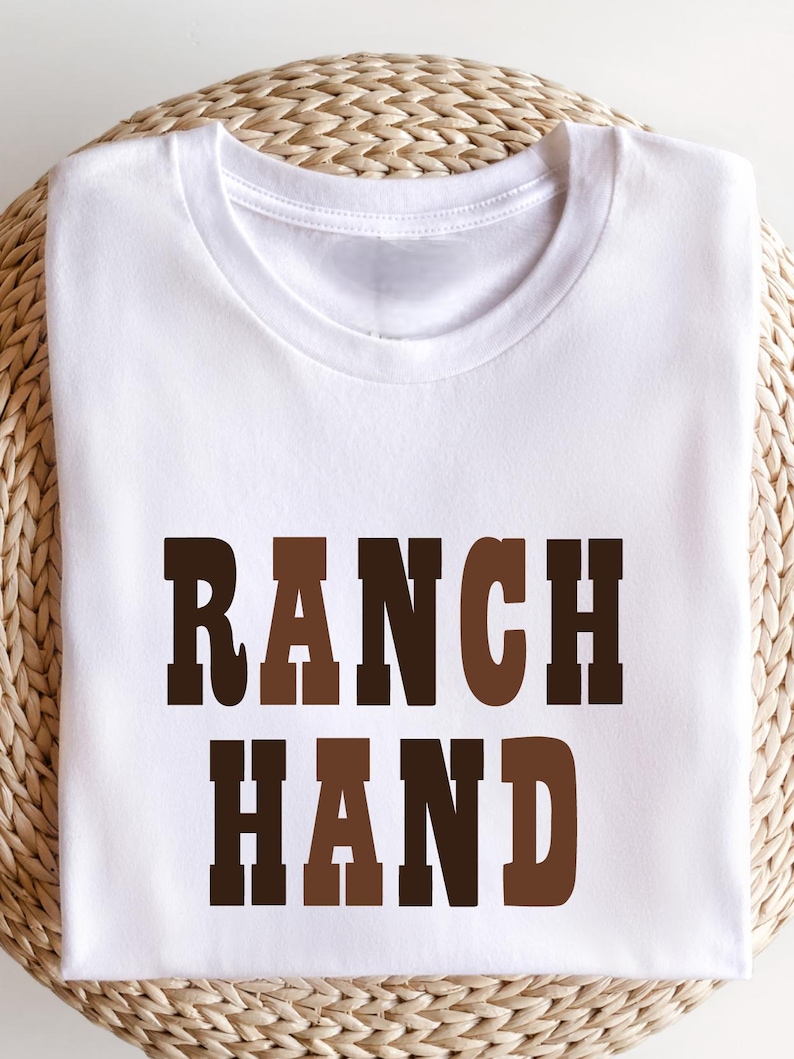 May include: A white t-shirt with the words "RANCH HAND" printed in brown and dark brown block letters. The shirt is folded and resting on a woven, light brown surface.