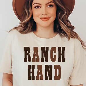 May include: A woman wearing a beige t-shirt with the words "RANCH HAND" in brown block letters. She is also wearing a brown hat. The image is well-lit and the woman is smiling.
