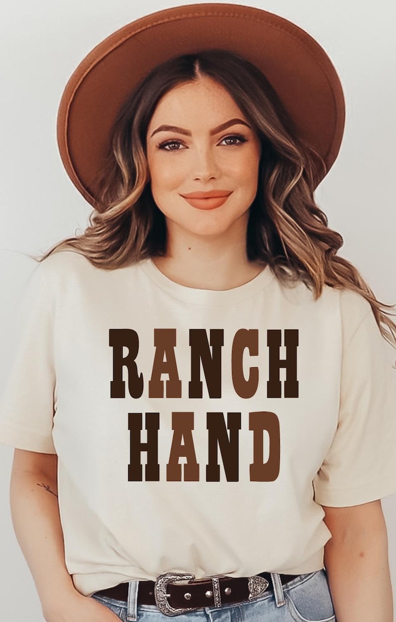 May include: A woman wearing a beige t-shirt with the words "RANCH HAND" in brown block letters. She is also wearing a brown hat. The image is well-lit and the woman is smiling.