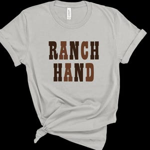 May include: A light gray t-shirt with the words "RANCH HAND" printed in brown block letters. The shirt is made of a soft material and has a crew neck and short sleeves. The shirt is knotted at the bottom.