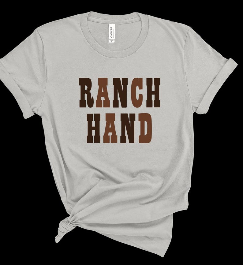May include: A light gray t-shirt with the words "RANCH HAND" printed in brown block letters. The shirt is made of a soft material and has a crew neck and short sleeves. The shirt is knotted at the bottom.