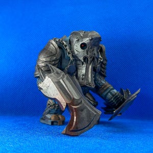 Könnte beinhalten: Detaillierte Miniaturfigur einer Fantasie-Kreatur, möglicherweise ein Krieger oder Monster. Die Figur ist hauptsächlich grau mit metallischen Akzenten, einschließlich Rüstung und einer großen Axt. Die Kreatur hat einen bedrohlichen Ausdruck mit einem roten Auge, vor blauem Hintergrund.