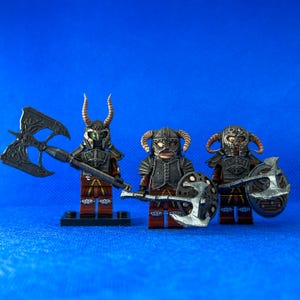 Könnte beinhalten: Drei maßgeschneiderte Lego-Minifiguren, die als Charaktere aus dem Videospiel Skyrim verkleidet sind. Die Figuren tragen dunkle Rüstungen mit Hörnern und halten Waffen. Die Figuren sind auf einem blauen Hintergrund platziert.