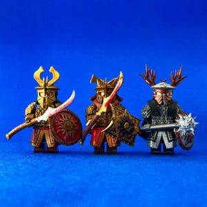 Könnte beinhalten: Drei Miniaturfiguren von Kriegern, die Rüstungen tragen und Waffen halten. Die Figuren sind aus Kunststoff und in Gold, Rot und Silber bemalt. Die Figur links hält ein Schwert und einen Schild, die Figur in der Mitte hält ein Schwert und einen Schild, und die Figur rechts hält eine Keule und einen Schild.