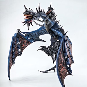 Könnte beinhalten: Detaillierte blaue Drachenfigur mit roten Akzenten auf den Flügeln und einem Reiter. Der Drache hat scharfe Zähne, Stacheln und ist angekettet. Die Flügel sind ausgebreitet, vor weißem Hintergrund.