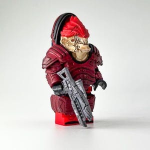 Könnte beinhalten: Eine rote und graue, benutzerdefinierte Lego-Minifigur eines fiktiven außerirdischen Kriegers, der eine futuristische Waffe hält. Die Figur hat eine detaillierte Kopfskulptur und Rüstung.