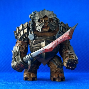 Könnte beinhalten: Eine braune und graue Actionfigur einer Kreatur mit einem großen, scharfen Schwert. Die Kreatur trägt einen Helm mit einem Kettenhemdkragen und hat einen grimmigen Ausdruck.