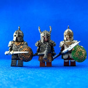 Könnte beinhalten: Drei silberne und goldene Miniaturfiguren von Kriegern mit Helmen, Schilden und Schwertern. Die Figuren stehen auf einem blauen Hintergrund.