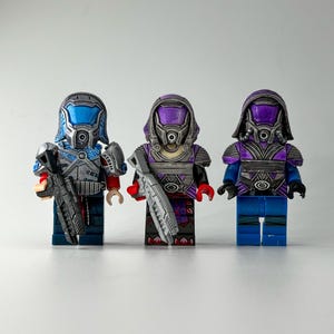 Könnte beinhalten: Drei individuell bemalte Lego-Minifiguren, jede mit einem Helm und einer Waffe. Die Minifiguren sind blau, lila und lila.