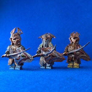 Könnte beinhalten: Drei maßgeschneiderte Lego-Minifiguren, die als Elfen verkleidet sind, jede mit einem einzigartigen Helm und einem Schwert mit einem dekorativen Griff. Die Figuren sind auf einem blauen Hintergrund platziert.