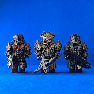 Puede incluir: Tres figuras de acción detalladas con armaduras ornamentadas en tonos marrón, bronce y plata, cada una con armas únicas. Las figuras están sobre una superficie azul, mostrando diseños intrincados.