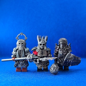 Könnte beinhalten: Drei detaillierte, silberfarbene, Miniaturfiguren von Kriegern mit Helmen und Waffen. Die Figuren stehen auf einer blauen Oberfläche.