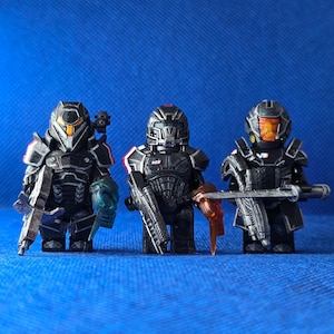 Peut inclure: Trois figurines miniatures en plastique vêtues d'armures tactiques noires avec des casques détaillés. Chaque figurine tient un accessoire d'arme unique, incluant des fusils et des lames. Les figurines présentent des accents orange et bleu translucide.