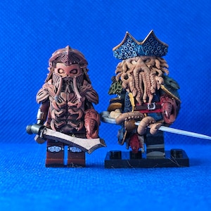 Könnte beinhalten: Zwei detaillierte Piratenfiguren. Eine Figur ist braun mit einem Schwert und klauenartigen Händen. Die andere Figur trägt einen blauen Hut und hat Tentakel als Bart und hält ein Schwert. Beide Figuren stehen auf schwarzen Sockeln vor blauem Hintergrund.
