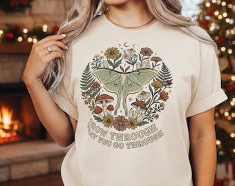 May include: Cream-colored t-shirt featuring a circular design with a green moth, mushrooms, flowers, and crystals. The text "GROW THROUGH WHAT YOU GO THROUGH" is arched around the bottom of the design. The shirt has short sleeves.