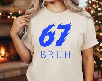 Può includere: Una maglietta color crema con il numero "67" in blu, stilizzato con bordi a forma di fiamma. Sotto il numero, la parola "BRUH" è stampata in blu. La maglietta ha maniche corte e girocollo.