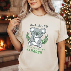 Può includere: T-shirt beige con la scritta "KOALAFied Human Resources MANAGER". La maglietta presenta un koala dei cartoni animati con gli occhiali e una penna, circondato da foglie verdi. Il design è in nero e verde.