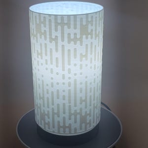 Puede incluir: Una lámpara de mesa cilíndrica con una pantalla blanca estampada. La pantalla presenta un diseño repetitivo de líneas verticales y puntos, e ilumina desde dentro. La lámpara se asienta sobre una base gris oscuro.