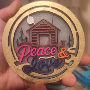 May include: A round wooden ornament with a gold border and a cabin scene inside. The cabin is brown with a white roof and a white moon above it. The words "Peace & Love" are written in a colorful script below the cabin.