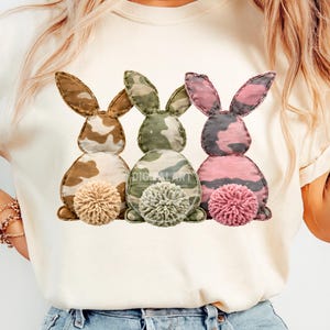 Könnte beinhalten: Cremefarbenes T-Shirt mit drei Hasen-Designs mit Tarnmustern in Braun, Grün und Rosa. Jeder Hase hat einen flauschigen Pompon-Schwanz. Der Text "DIGITAL ART" ist auf dem Shirt zu sehen.