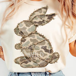 以下が含まれることがあります： クリーム色のTシャツに迷彩柄のウサギのデザイン。ウサギは緑、茶色、タン色の布のパッチでできており、糸が見えるように縫い付けられています。「DIGITAL ART WILD」の文字がデザインの一部に表示されています。