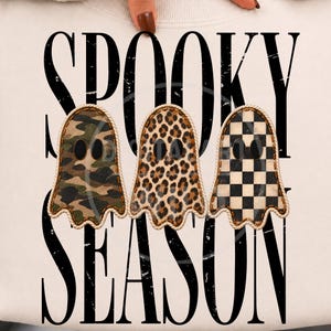 Può includere: Una felpa di colore chiaro con la scritta nera invecchiata SPOOKY SEASON. Tre toppe a forma di fantasma sono centrate, ognuna bordata da una corda. I fantasmi presentano motivi mimetici, leopardati e a scacchi bianchi e neri.