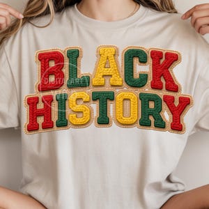 May include: A cream-colored t-shirt featuring the words "BLACK HISTORY" in a bold, textured design. The text is in red, yellow, and green, with a stitched appearance, and a tan border. The shirt is a casual style.