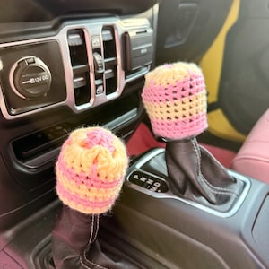 May include: Two crochet covers for vehicle gear shifters, each featuring alternating horizontal stripes of pale yellow and light pink yarn. The covers are fitted over the top of the black leather gear shift knobs in a vehicle center console.