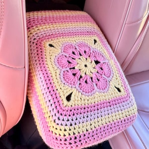 May include: A handmade crochet car seat cover in shades of pink and yellow. The square cover features a central floral design and striped borders. The cover is placed in a pink car interior, adding a touch of handmade charm.